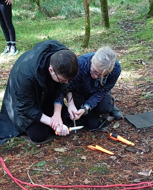 Two people kneeling in a forest, working together with tools on the ground, surrounded by green trees Two people kneeling in a forest, working together with tools on the ground, surrounded by green trees